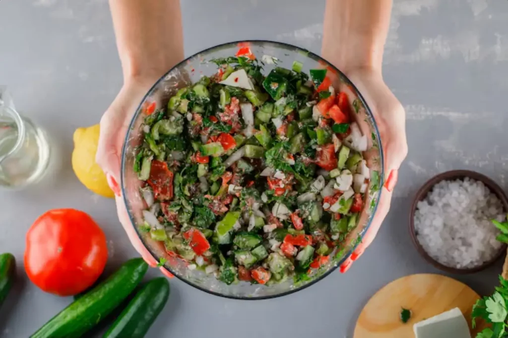 Woman holding vegetable salad with tomatoes, cheese, cucumber, salt in a glass bowl on gray surface, top view