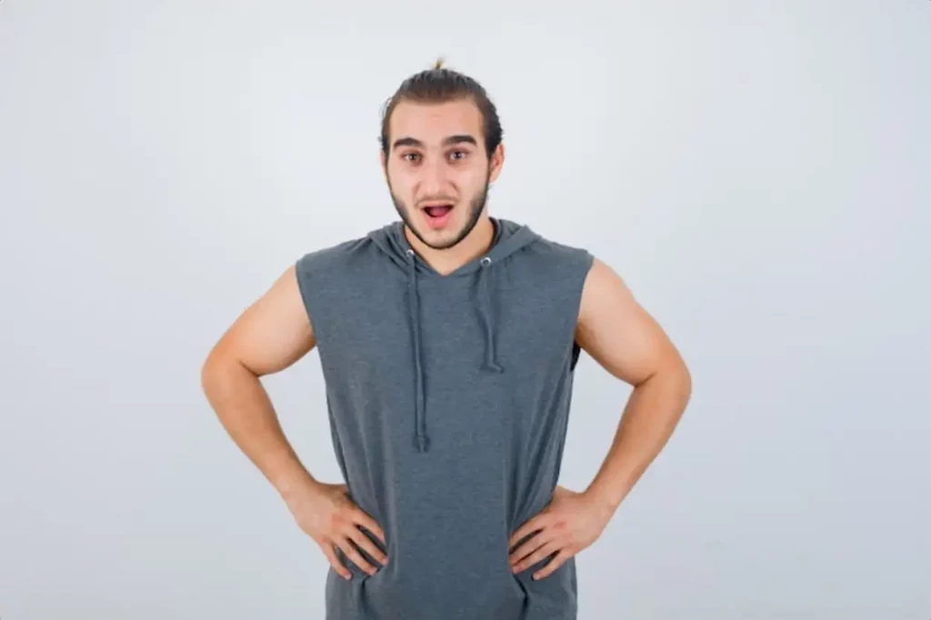 Young fit male posing with hands on waist in sleeveless hoodie and looking shocked , front view.