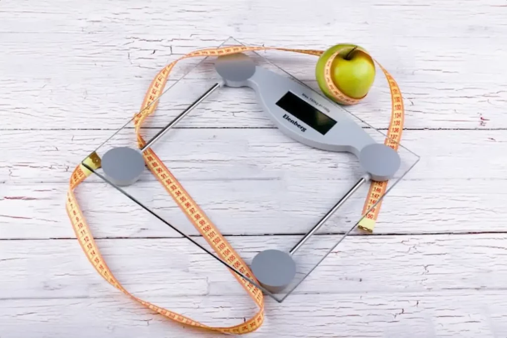 Green apple, orange tape-measure lie on glass weighing scales